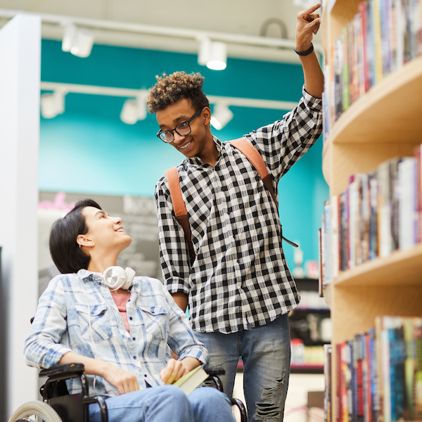 Dos personas hacen compras juntas en una librer&iacute;a. Una est&aacute; en una silla de ruedas y la otra se&ntilde;ala un libro en una repisa.